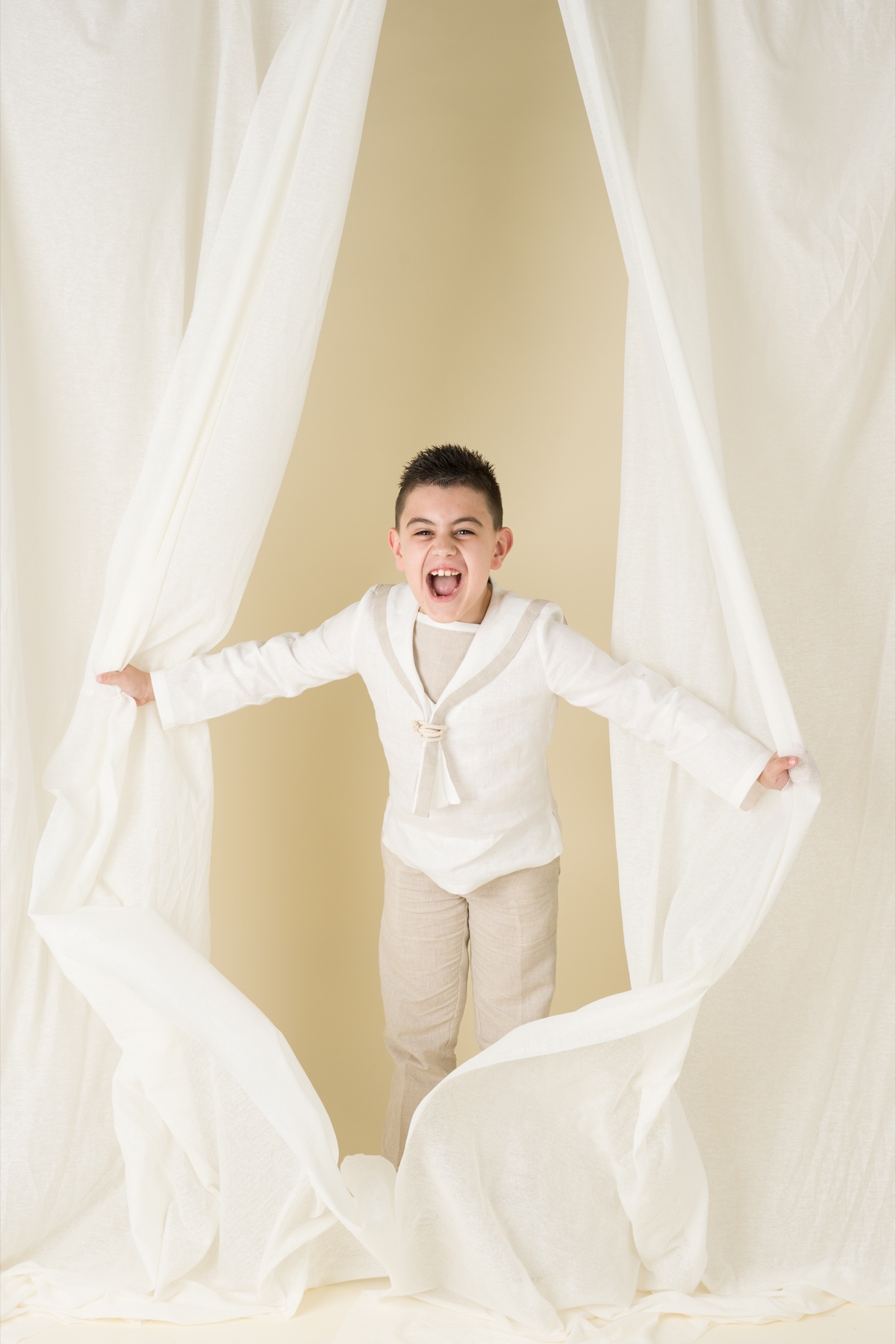Niño con traje de comunión abriendo grandes cortinas blancas.