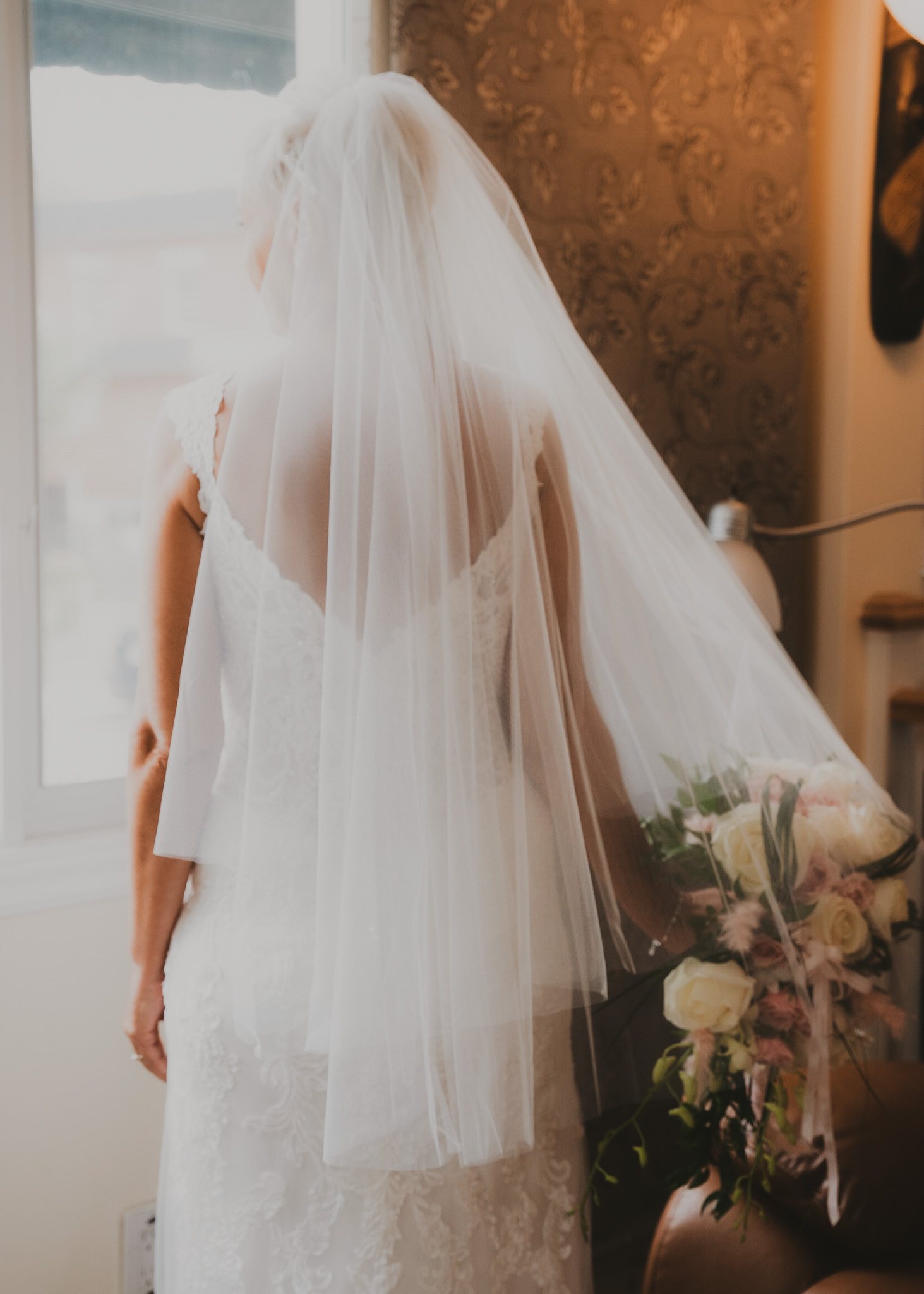 Le dos et le voile d'une mariée devant une fenêtre lumineuse.